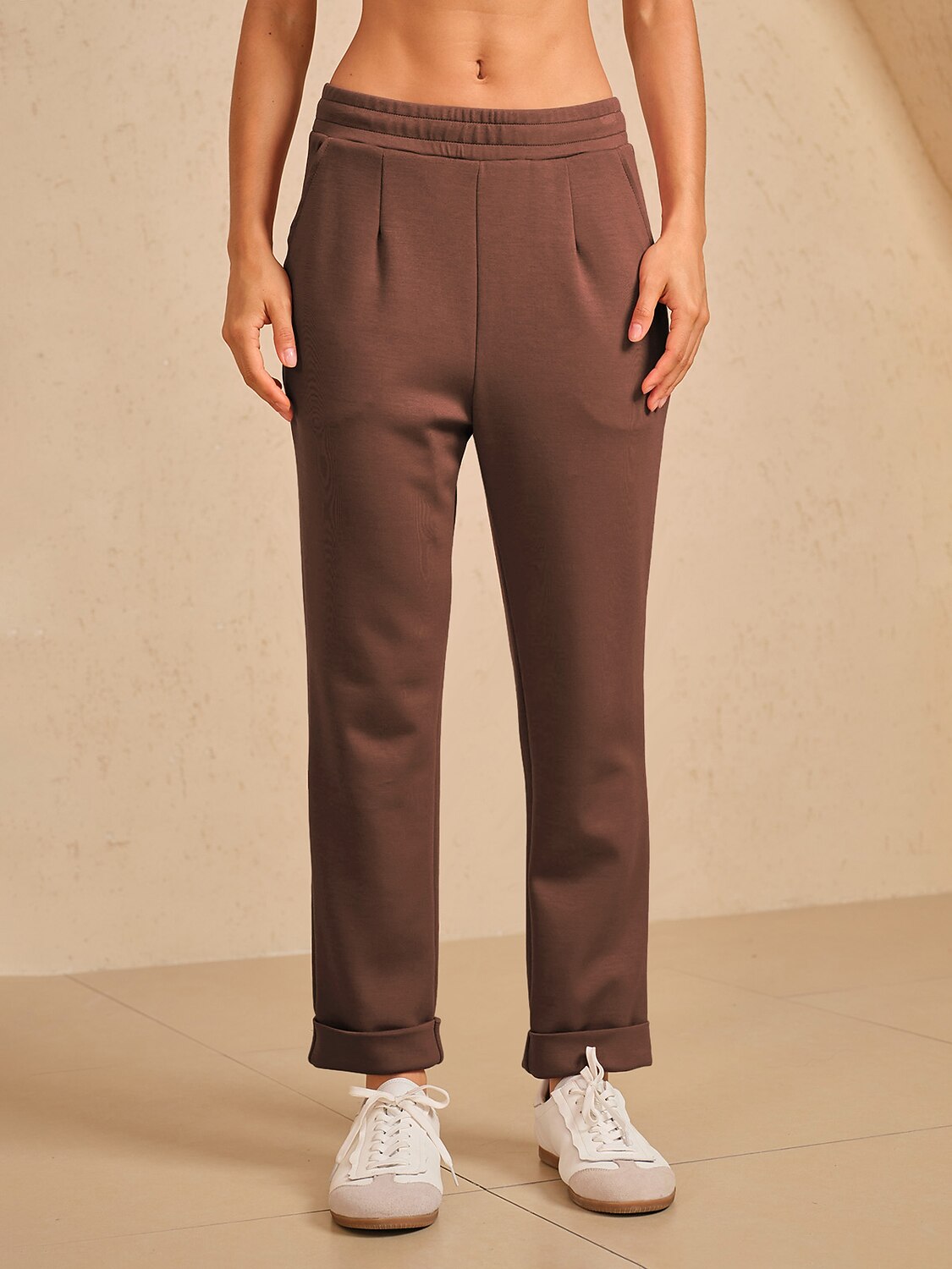 Woman wearing brown high-waisted pleated trousers and white sneakers.