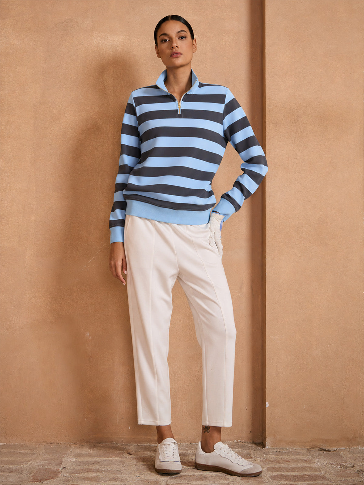 Woman wearing a blue and grey striped quarter-zip golf pullover and white performance trousers.
