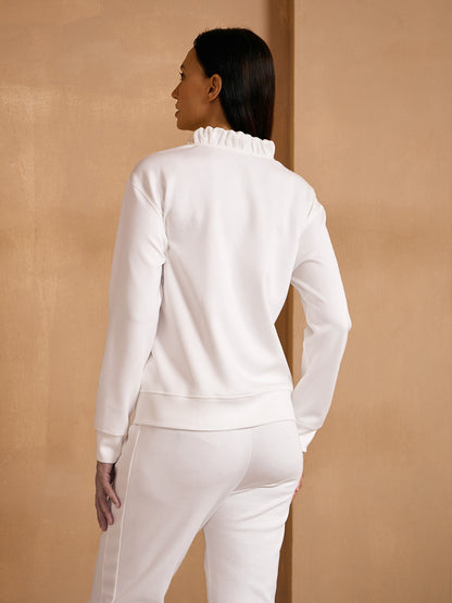Woman wearing a white long-sleeve ruffled collar sweatshirt and matching white trousers.